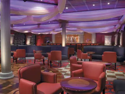 Panoramablick in eine stilvolle Bar-Lounge auf der Carnival Freedom. Rote Ledersessel, dunkle Sofas, ein großer Barbereich mit Statuen und eine Decke mit lila beleuchteten Stoffdrapierungen sind zu sehen.