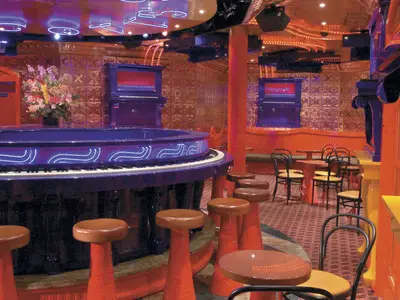 Eine luxuriöse Bar-Lounge auf der Carnival Freedom mit einer auffälligen runden, violetten Klavierbar im Vordergrund, um die sich Barhocker reihen. Im Hintergrund kleine Tische und Stühle vor goldgeprägten Wänden und orangefarbenen Säulen, die eine warme und einladende Atmosphäre schaffen.