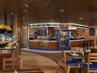 Innenansicht des mexikanischen Buffetrestaurants 'Blue Iguana Cantina' auf der Carnival Freedom. Mehrere Selbstbedienungsstationen mit frischen Zutaten für Tacos und Burritos sind zu sehen, sowie Tische und Stühle für Gäste.