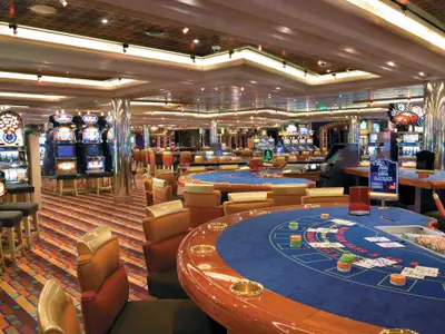 Ein weitraeumiges, helles Casino auf der Carnival Freedom, ausgestattet mit zahlreichen bunten Spielautomaten, runden Blackjack-Tischen mit blauer Filzoberfläche und bequemen Stühlen in warmen Farben. Keine Personen sichtbar.