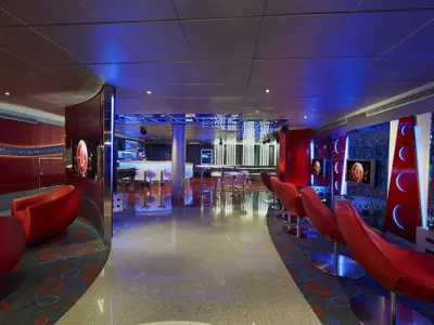 Ein moderner Club auf der Carnival Freedom mit roter und blauer LED-Beleuchtung. Bequeme rote Sessel stehen vor Bildschirmen. Im Hintergrund eine Bar mit hohen Hockern und ein Kicker-Tisch. Der Boden ist glänzend.
