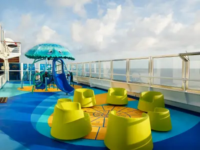 Ein farbenfroher Kinderbereich auf der Carnival Freedom mit Rutsche, pilzförmigem Dach und hellgrünen Sitzen. Das Deck hat ein blau-orangefarbenes Muster mit Zahlen. Im Hintergrund ist das Meer unter einem bewölkten Himmel zu sehen.