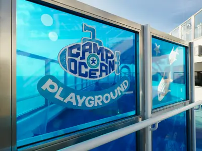 Blau getöntes Fenster auf der Carnival Freedom mit 'Camp Ocean Playground' Logo, U-Boot und Fischen. Im Hintergrund Himmel und Schiffsdeck.