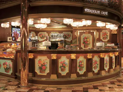 Ein üppig eingerichtetes Wiener Café an Bord der Carnival Freedom mit einem Tresen voller Kuchen und Desserts. Goldene Akzente und florale Gemälde schmücken den Raum, dazu Kristallleuchter und eine Kaffeemaschine.