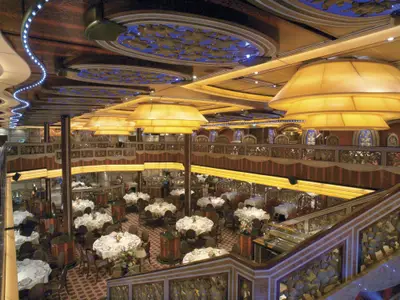 Ansicht eines großen, zweistoeckigen Restaurants auf der Carnival Freedom. Mehrere runde Tische mit weißen Tischdecken und Besteck sind zu sehen. Die Decke ist mit blauen, beleuchteten Paneelen und langen, gelben Stofflampen verziert. Aufwendige Holzgeländer und Wanddekorationen prägen den Raum.