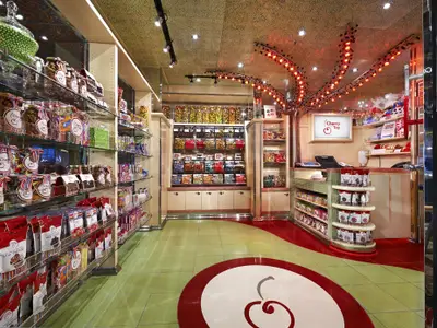 Innenansicht eines farbenfrohen Süßigkeitenladens an Bord. Regale sind mit Gläsern, Tüten und Boxen voller Bonbons und Schokolade gefüllt. Ein 'Cherry Top'-Schild leuchtet über der Kasse, der Boden ist grün und rot mit einem Kirschenmotiv.