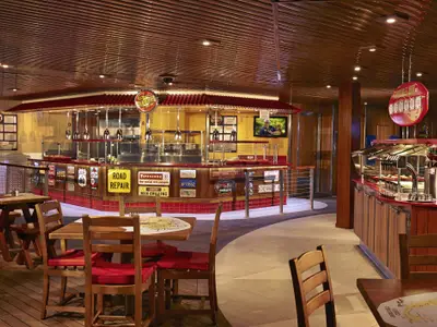 Eine weite Innenansicht eines rustikalen Burger-Restaurants an Bord, dekoriert mit Nummernschildern und Straßenschildern. Zu sehen sind eine lange Theke für Burger und eine separate Theke für Beilagen und Saucen, sowie mehrere Tische mit Holzstühlen und roten Kissen.