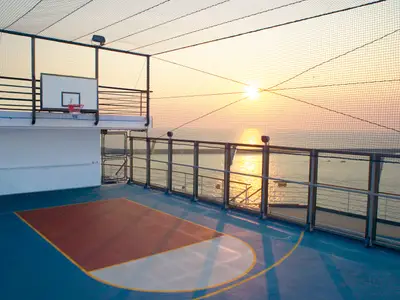 Basketballfeld auf dem Oberdeck der Carnival Freedom bei Sonnenuntergang. Links der Korb, rechts das Meer mit der tiefstehenden Sonne. Ein Schutznetz umgibt den Platz.