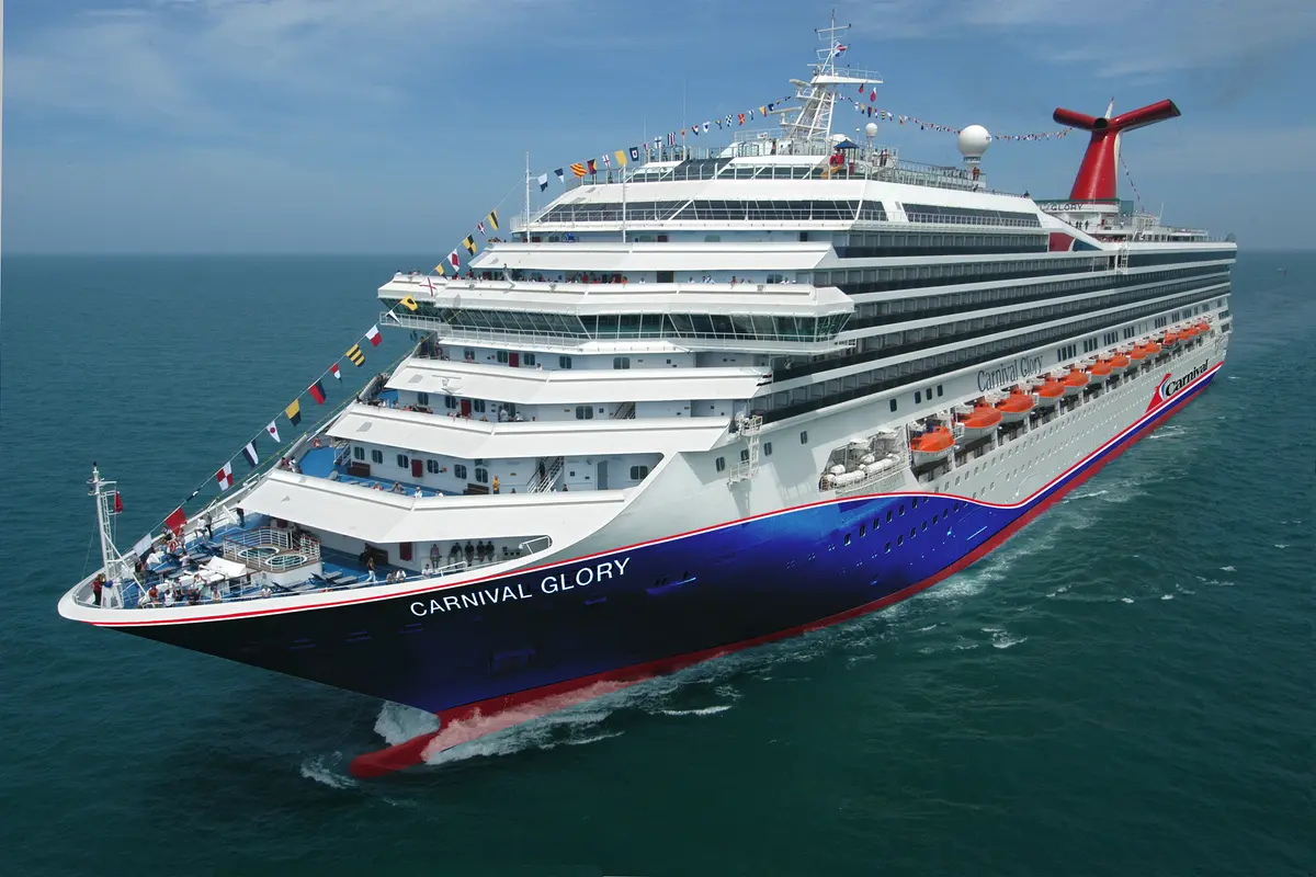 Die Carnival Glory, dieses große Schiff mit blau-weißem Rumpf und rotem Schornstein, fährt auf blauem Meer. Bunte Flaggen schmücken den Bug, Passagiere sind auf den Decks sichtbar.