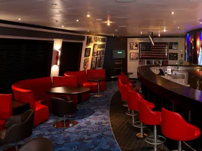 Eine elegant gestaltete Bar-Lounge an Bord der Carnival Glory. Die Einrichtung umfasst eine geschwungene Bartheke mit roten Barhockern und mehrere rote Sofas und Tische. An den Wänden hängen gerahmte Bilder und eine große amerikanische Flagge. Ein großer Bildschirm zeigt eine Bühnenperformance.