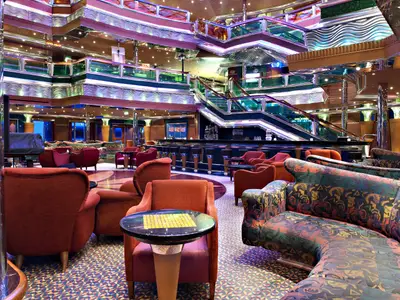Geräumige, mehrstöckige Lounge auf der Carnival Glory mit einer langen Bar. Im Vordergrund sind zahlreiche rote und gemusterte Sessel und Tische zu sehen. Im Hintergrund führen breite Treppen in höhere Decks. Die Decken und Wände sind mit glänzenden, reflektierenden Materialien und farbiger Beleuchtung dekoriert.
