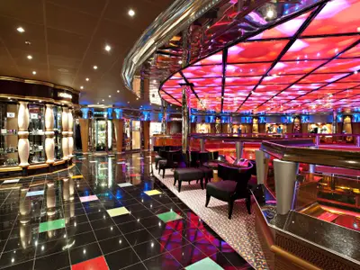 Ein lebhaft beleuchteter Innenbereich der Carnival Glory mit einer modernen Bar-Lounge. Links sind gläserne Schaufenster von Geschäften zu sehen. Der Boden ist schwarz glänzend mit farbigen Quadraten, während die Decke leuchtend rote Paneele und Akzentbeleuchtung aufweist. Schwarze Sessel und Tische stehen im Loungebereich.