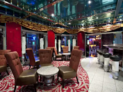 Innenansicht einer opulenten Schiffsbar mit roten Akzentwänden, gemustertem Teppich, Spiegeldecken und Barhockern. Verschiedene Sitzecken mit Tischen und gepolsterten Stühlen sind zu sehen, sowie dekorative Kerzensäulen.