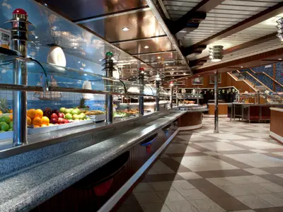Das helle Buffet-Restaurant der Carnival Glory zeigt eine lange Theke mit frischem Obst, wie Äpfeln und Orangen, sowie weiteren Speisen. Im Hintergrund eine maritime Wandbemalung mit Segelschiffen. Schachbrettartiger Boden.