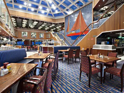 Ein helles und geräumiges Buffetrestaurant auf der Carnival Glory mit Holzakzenten, blauen Polstermöbeln, Esstischen, einem Buffetbereich und einer großen Segelboot-Skulptur an der Wand.