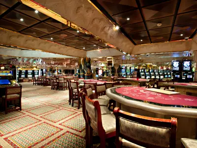 Ein leeres Casino auf der Carnival Glory mit vielen Reihen von Spielautomaten und mehreren roten Spieltischen. Die Decke ist reich verziert im altägyptischen Stil mit Skulpturen und goldenen Akzenten, darunter Anubis-Statuen.