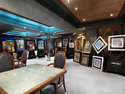 Geräumige Kunstgalerie auf der Carnival Glory. Zahlreiche moderne Gemälde in Rahmen sind auf Staffeleien ausgestellt. Helle blaue Deckenbeleuchtung und bequeme Holzstühle mit gestreiften Polstern laden zum Verweilen ein.