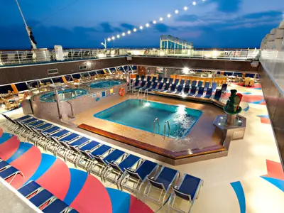 Blick von oben auf den beleuchteten Poolbereich der Carnival Glory bei Abenddämmerung. Ein großer Pool, zwei Whirlpools, viele leere Liegestühle, Tische und Stühle sind zu sehen. Eine Lichtgirlande spannt sich über das Deck.