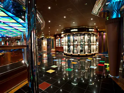 Ein moderner Einkaufsbereich auf der Carnival Glory. Der Raum hat einen glänzenden schwarzen Fliesenboden mit bunten Quadraten. Die Decke ist mit farbigen, geometrischen Lichtpaneelen ausgestattet. Elegante Vitrinen zeigen Produkte. Eine helle, einladende Atmosphäre.