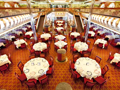 Ein großer, eleganter Speisesaal auf der Carnival Glory, gesehen aus der Vogelperspektive. Mehrere weiß gedeckte Tische mit roten Stühlen stehen auf einem gemusterten Teppich. Eine prächtige Treppe führt zu einer oberen Ebene mit weiteren Tischen und Sitznischen.