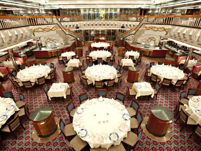 Weitwinkelaufnahme eines eleganten, mehrstöckigen Restaurants auf der Carnival Glory mit zahlreichen festlich gedeckten Tischen. Eine imposante Treppe mit kunstvollen Wandgemälden und Dekorationen flankiert den Speisesaal.