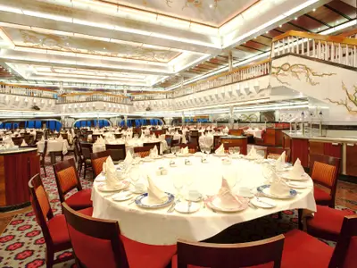 Geräumiger, zweistöckiger Speisesaal der Carnival Glory mit zahlreichen festlich eingedeckten Tischen und einer kunstvoll bemalten Treppe.