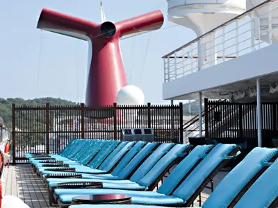 Reihe blauer Liegestühle auf einem Sonnendeck der Carnival Glory. Man sieht den ikonischen roten Schornstein, einen weißen Radom und eine bewaldete Küstenlinie.