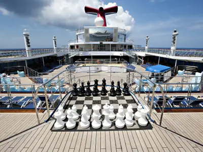 Blick auf das obere Sonnendeck der Carnival Glory mit einem riesigen Schachspiel im Vordergrund, Lounge-Bereichen und einem großen Bildschirm im Hintergrund. Das Meer ist am Horizont sichtbar unter blauem Himmel mit Wolken.