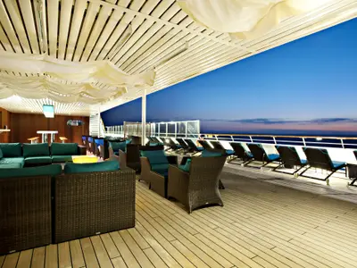 Abendliche Szene auf dem Sonnendeck der Carnival Glory mit Rattan-Sitzgruppen, grünen Polstern und zahlreichen Liegestühlen entlang der Reling. Im Hintergrund der blaue Himmel und das Meer bei Dämmerung.
