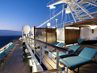 Abendliche Szene auf dem Oberdeck der Carnival Glory. Türkisblaue Liegestühle und Loungemöbel stehen auf dem Holzdeck. Im Hintergrund ist das offene Meer und ein dunkler Himmel zu sehen. Weiße Radarkuppeln und eine beleuchtete Segellinie tragen zur Atmosphäre bei. Eine Person entspannt in einer Hängeliege unter einem Holzpavillon.