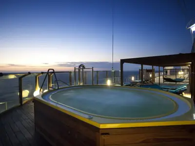 Blick auf einen beleuchteten Whirlpool auf der Carnival Glory bei Sonnenuntergang. Im Hintergrund das Meer und ein dunkler Himmel. Liegestühle und eine Hängematte unter einer Pergola sind sichtbar. Die Szene ist ruhig und entspannend.