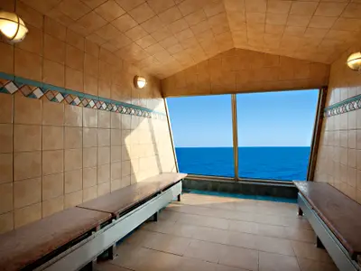 Ein gefliester Innenraum auf der Carnival Glory mit langen Holzbänken an den Seiten. Eine breite Fensterfront bietet einen weiten Blick auf das tiefblaue Meer und den Horizont unter einem klaren blauen Himmel. Die Wände sind in warmen Brauntönen gefliest und mit einer dekorativen Bordüre versehen. Zwei Lampen sind an den Seitenwänden sichtbar. Die Szene ist hell beleuchtet.