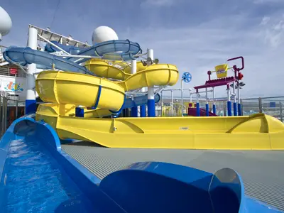 Ein lebhafter Wasserpark auf dem Deck der Carnival Glory, mit gelben und blauen Rutschen, einem großen Kippeimer und Wasserspielen unter blauem Himmel.