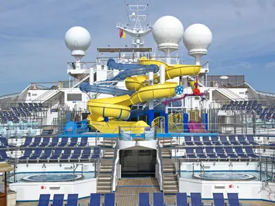 Sonniges Deck der Carnival Glory mit auffälligen gelben und blauen Wasserrutschen, Liegestühlen und Whirlpools. Im Hintergrund ragen Radarkuppeln in den blauen Himmel.