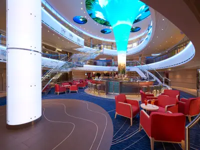 Modernes, helles Atrium auf der Carnival Horizon. Eine zentrale Bar, rote Sessel und eine leuchtende Säule mit digitalen Anzeigen in der Mitte. Runde Deckenbildschirme sind ebenfalls zu sehen.
