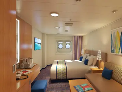 Geräumige und hell beleuchtete Außenkabine auf der Carnival Horizon. Die Kabine verfügt über ein Doppelbett, ein Sofa, einen Tisch und zwei runde Portholes mit Blick auf das Meer. Ein Fernseher und ein Kunstwerk mit bunten Surfbrettern schmücken die Wände.