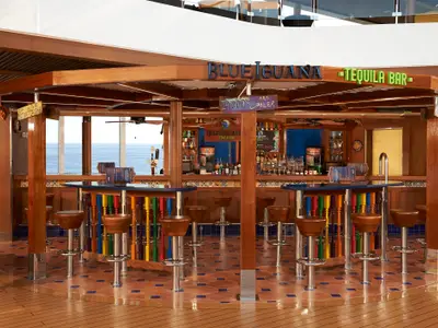 Eine farbenfrohe Blue Iguana Tequila Bar auf der Carnival Horizon mit braunen Hockern, einer blauen Theke und weitem Blick auf das offene Meer durch große Fenster. Die Bar hat Holzverkleidung und bunte Dekorationen.