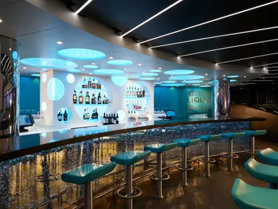 Eine futuristische Bar-Lounge mit einer langen, geschwungenen Theke, die mit unzähligen Lichtern beleuchtet ist. Türkisfarbene Barhocker stehen davor. Im Hintergrund sind Regale mit Spirituosen in runden Nischen zu sehen, alles in einem modernen, hellblauen Design.