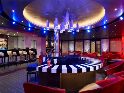 Eine moderne Bar-Lounge mit einem schwarzen Flügel, der eine weiß-schwarz gestreifte Bar umgibt. Rote Samtsessel sind um kleine Tische gruppiert. Links befindet sich eine lange Bar mit Barhockern. Die Decke ist mit blauen Lichtern und einer kunstvollen Kristallleuchte versehen. Zwei Cocktails stehen auf der Piano-Bar.
