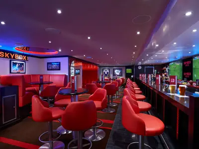 Moderne Sportsbar 'Skybox' an Bord. Rote Polstersitze, Tische und eine lange Theke mit Bier. Große Videowand zeigt Fussballspiel. Sporttrikots und Bilder an den Wänden. Lebendige Neonbeleuchtung.