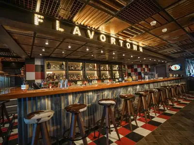 Eine Bar namens 'FLAVORTOWN' an Bord der Carnival Horizon, gekennzeichnet durch eine lange Holztheke, dahinter beleuchtete Regale voller Flaschen. Der Boden ist kariert in Rot, Schwarz und Weiß. Die Decke hat ein rustikales Design aus Metallplatten. Zahlreiche Hocker mit Kuhfellbezug stehen vor der Theke.