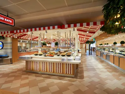 Eine Weitwinkelaufnahme eines hell erleuchteten Buffet-Restaurants auf der Carnival Horizon. Es sind lange Essensstationen mit verschiedenen Speisen und Tellern sowie ein "Beer Station"-Schild zu sehen. Die Decke ist mit rot-weißen Markisenstreifen verziert.