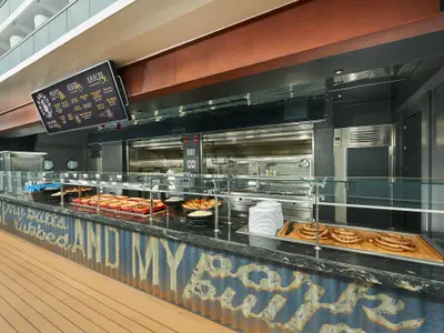 Eine Selbstbedienungstheke an Bord der Carnival Horizon mit einer Auswahl an warmen Speisen, Wurst und Beilagen. Im Hintergrund ist die offene Küche sichtbar, im Vordergrund ein dekorativer Tresen.