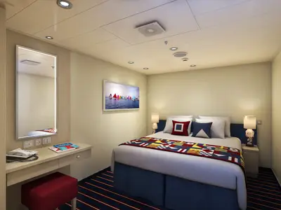 Blick in eine moderne Kreuzfahrtkabine mit Doppelbett, blauer Bettdecke und einem rot-blau-gelben Tagesläufer. An der Wand hängt ein Bild von Segelbooten. Links befindet sich ein Schreibtisch mit Telefon und Spiegel.
