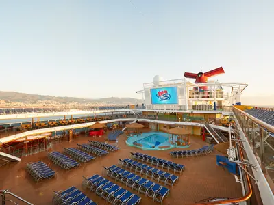 Hochwinkelansicht des Pooldecks der Carnival Horizon am Morgen, mit einem großen Pool, blauen Liegestühlen und einer riesigen LED-Leinwand, die 'Dive-In Movies' anzeigt. Im Hintergrund sind eine Küstenstadt und Berge zu sehen.