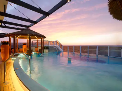 Ein luxuriöses Pooldeck auf der Carnival Horizon bei Sonnenuntergang. Das Hauptbecken und ein Whirlpool sind von Holzdecks umgeben. Ein schilfgedeckter Pavillon spendet Schatten, während orangefarbene Blumentöpfe dekorativ platziert sind. Der Himmel zeigt leuchtende Rosa- und Orangetöne.