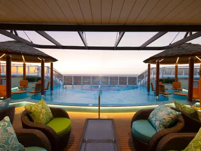 Ein luxuriöser Poolbereich auf der Carnival Horizon bei Sonnenuntergang. Der Pool ist von komfortablen Lounge-Sesseln und Tiki-Bar-ähnlichen Überdachungen umgeben. Im Hintergrund erstreckt sich das weite Meer.