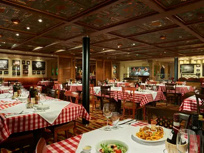 Innenansicht eines italienischen Restaurants an Bord der Carnival Horizon. Zahlreiche Tische mit rot-weiß karierten Tischdecken und gedeckten Tellern, Gläsern und Weinflaschen. Im Hintergrund ist eine offene Küche mit Köchen zu sehen. Die Decke ist mit kunstvollen Kupferplatten verziert.