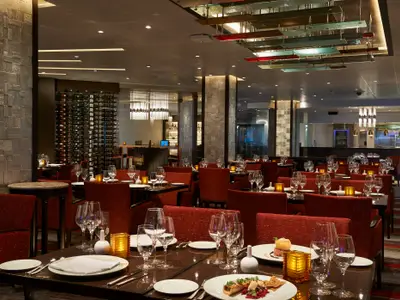 Ein elegantes Restaurant mit modernem Interieur. Rote Polsterstühle und dunkle Tische sind eingedeckt. Ein großes Weinregal und eine Bar sind im Hintergrund sichtbar.