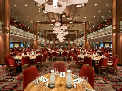 Großes, elegantes Restaurant der Carnival Horizon. Der Hauptbereich zeigt zahlreiche gedeckte Tische mit roten Stühlen. Im Hintergrund ist eine Bar zu sehen, und an den Seiten erstreckt sich eine obere Ebene mit weiteren Sitzgelegenheiten. Die Decke zieren moderne, quadratische Lichtinstallationen. Warme Holztöne dominieren das Interieur.